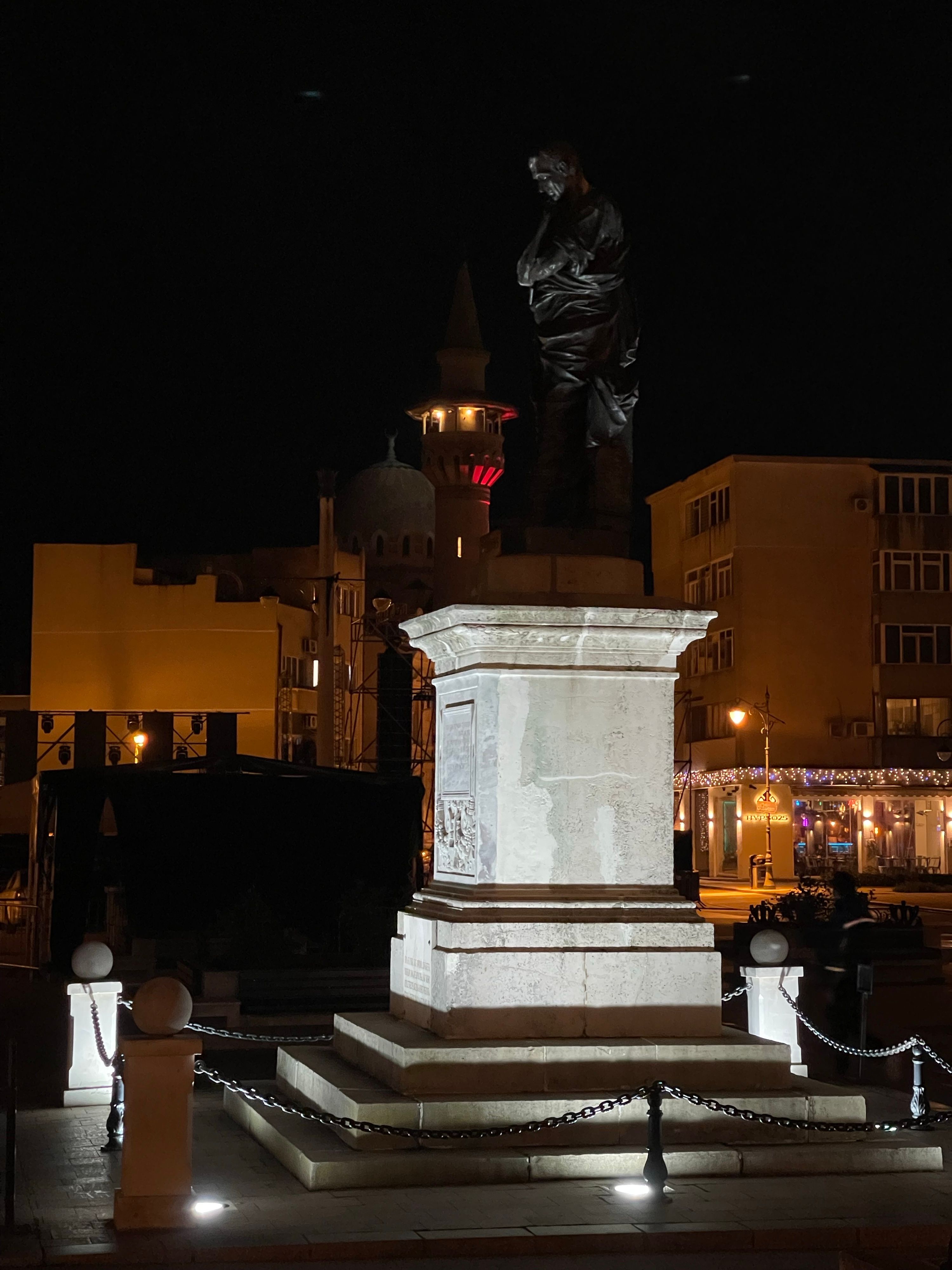 A nighttime photo of the statue of Ovid in Piața Ovidiu, Constanța, Romania, May 2026. The Grand Mosque and submarine steampunk bar Hypso25 are lit up in the background.