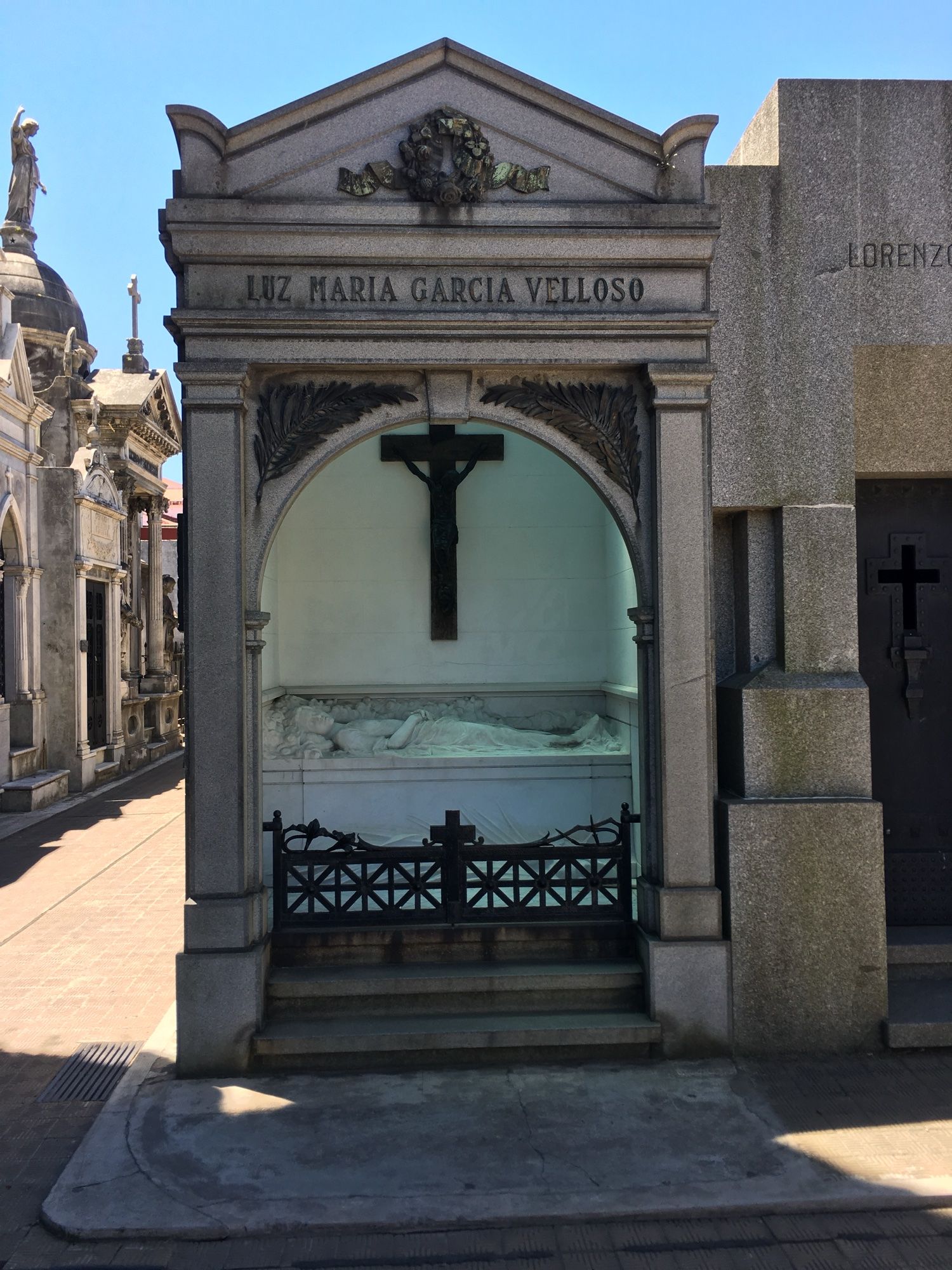 photo of an above ground, crypt-like grave, with an open arched entry and a white marble sculpture of a young woman in repose beneath a black cross.